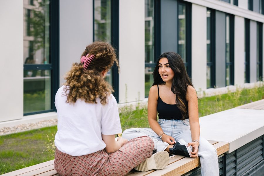 Zwei Studentinnen im Gespräch auf einer Bank im Außenbereich des bbw Campus